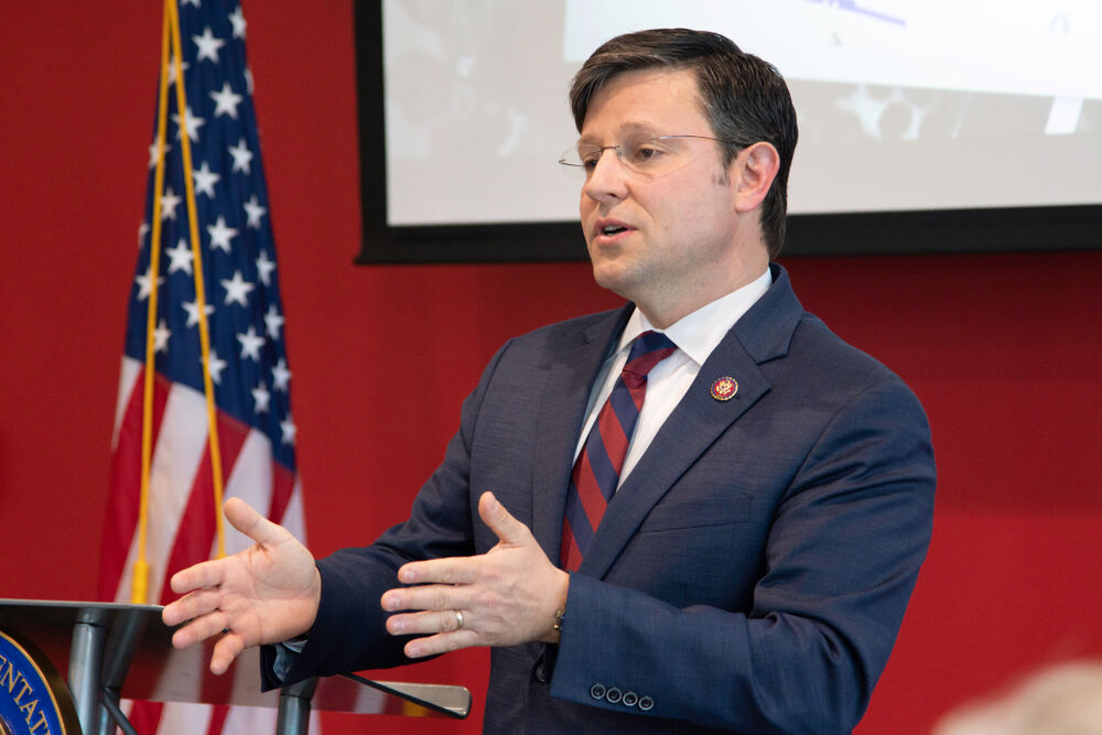 House Speaker Mike Johnson, a white brunette guy in a suit, gestures with his hands in a red room in front of an American flag and a display screen above him.