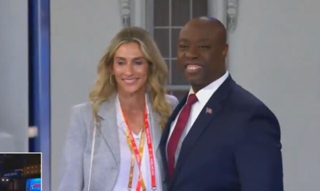 Tim Scott, a Black man in a blue suit with a red tie, stands next to a blonde white woman who is waring a grey blazer and an orange lanyard