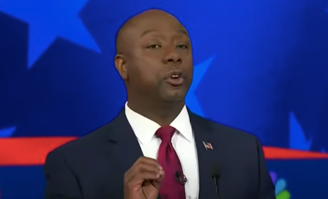 Tim Scott, a Black man in a dark suit and red tie, gestures his right hand while standing onstage with a blue background with stars on it.