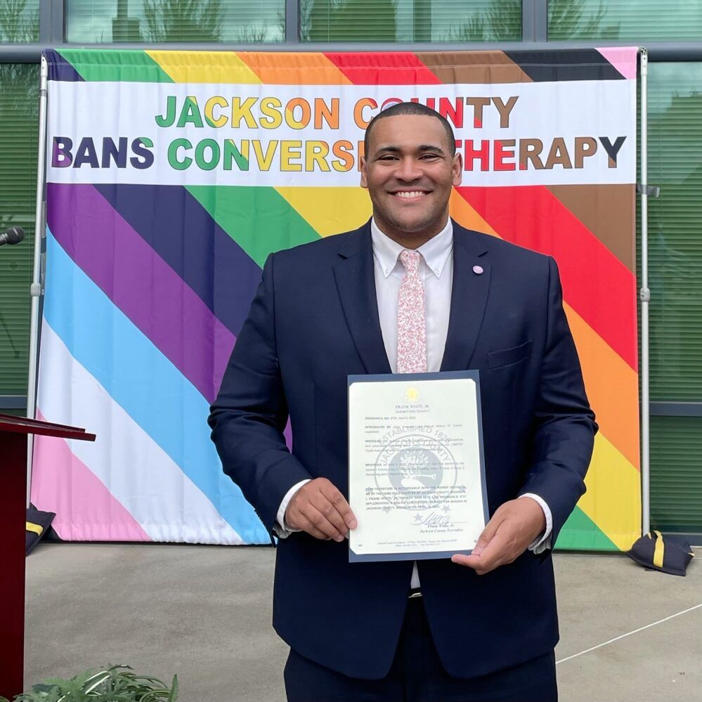 Justice Horn stands in front of a Progress Pride flag. He is smiling while holding a copy of the conversion therapy ban. 