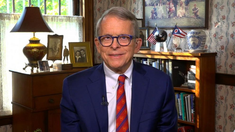 Ohio Gov. Mike DeWine is an old white guy. In this pic, he's wearing a red striped tie, a blue blazer, and glasses. He smiles whole sitting in a daylit office with state flags and framed pictures on bookshelves behind him.
