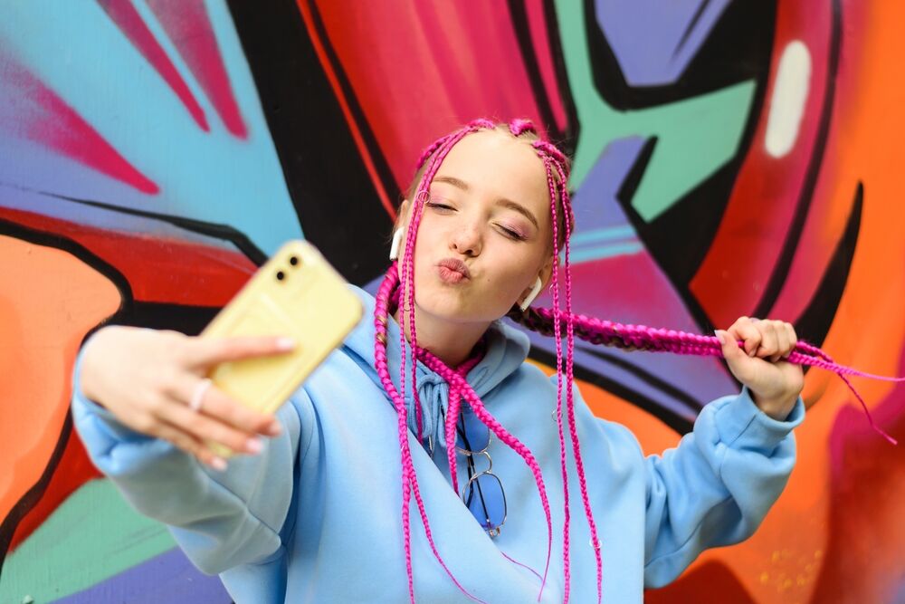 Caucasian teenage girl with pink braids is using a smartphone against the background of a multicolored street wall.