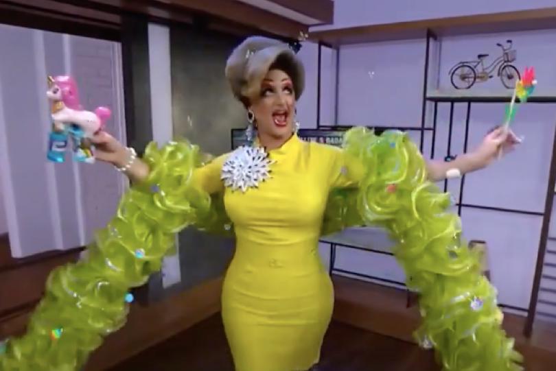 A drag queen with a coiffed blonde wig, a taffeta tufted boa, and a tight-fitting yellow dress accentuating her bosom and hips holds a unicorn-shaped bubble gun and a rainbow-colored pinwheel while dancing around a living room.
