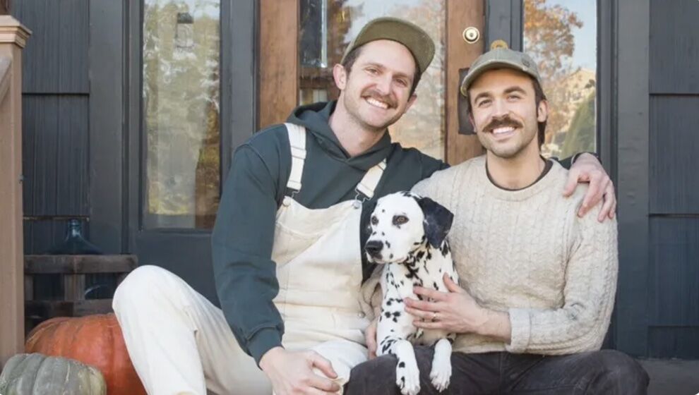 Jacob A. Carter (left) and Daniel Blagovich, are a married gay couple. They're both young, white, brunette men with moustachs. In this picture, they smile on he front steps of a house or business while holding their Dalmatian dog)