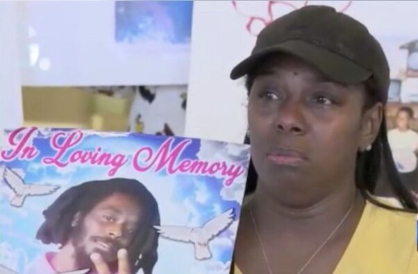 Marcia Irving, a Black woman holds a sign of her deceased son, Janard Geffrard. The sign says "In Loving Memory" in pink cursive and has a background of blue clouds