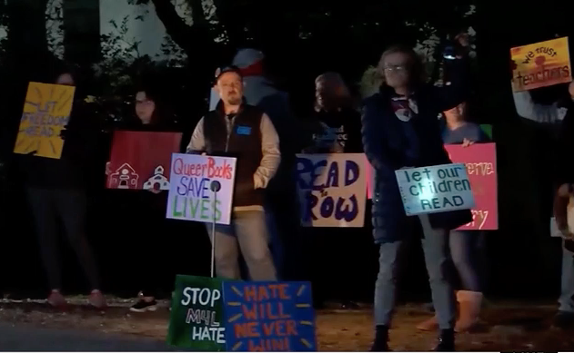 At night upon a sidewalk, protestors against Moms for Liberty carry hand-painted signs that say "Stop M4L hate," "Queer books save lives," "Hate will never win," "Let freedom read," "We trust teachers," and "We trust teachers."