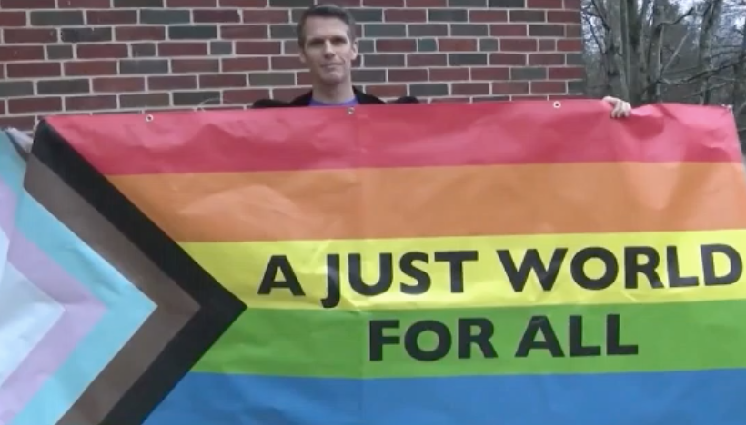 Pastor Matt Laney, a white grey-haired man of the cloth at the Virginia-Highland Church, holds the torn-down banner which is a rainbow progress Pride flag with the words "A just world for all" written on it.