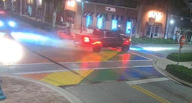A black truck with a flag flying from its back end screeches its tires, leaving black marks and smoke upon the rainbow crosswalk.