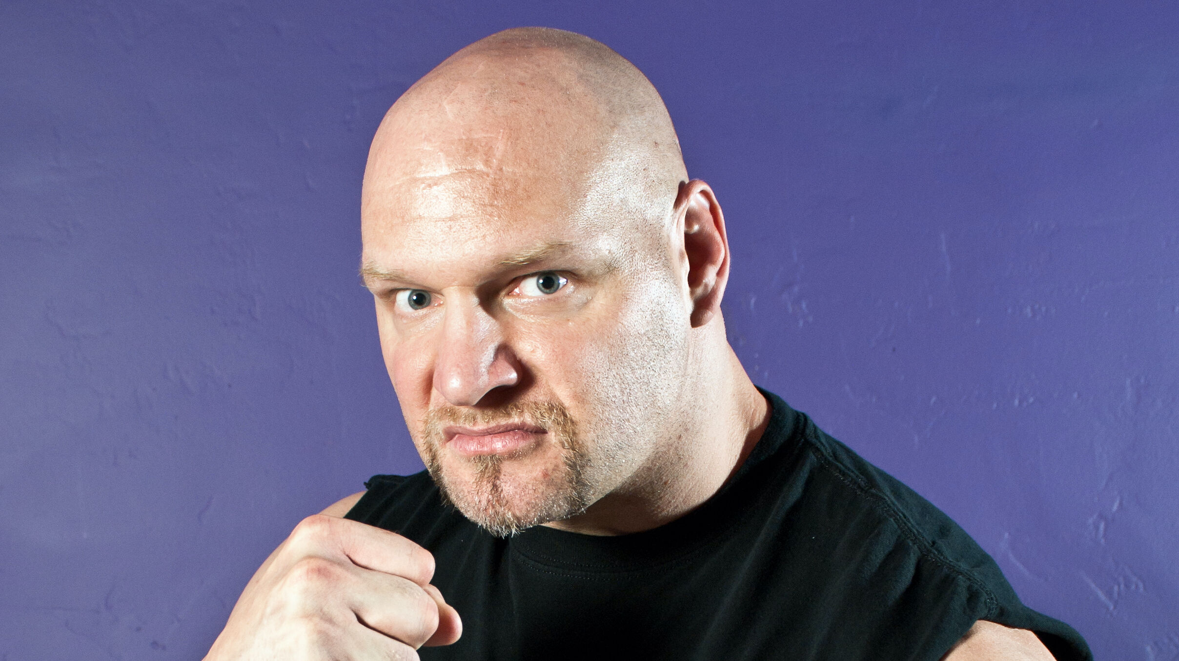 Val Venis is a bald white guy with a goatee. In this pic, he makes a fist and wears a black t-shirt while sneering against a purple background