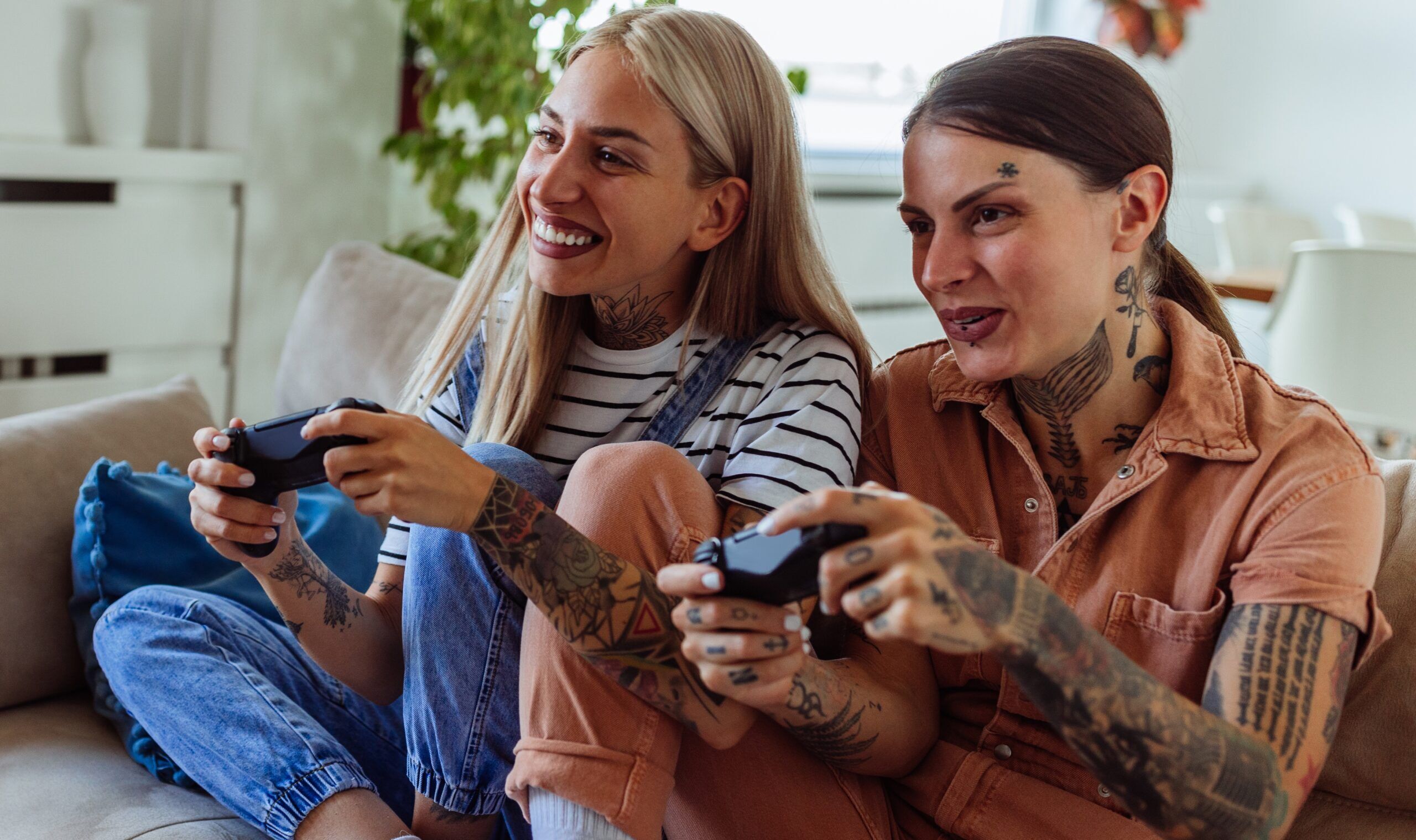 Cheerful lesbian couple playing video games together while being seated on a sofa at home during the day