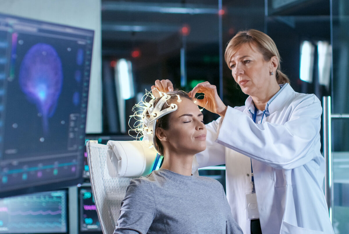 Woman Wearing Brainwave Scanning Headset Sits in a Chair while Scientist Adjusts the Device, Looks at Displays. In the Modern Brain Study Laboratory Monitors Show EEG Reading and Brain Model.