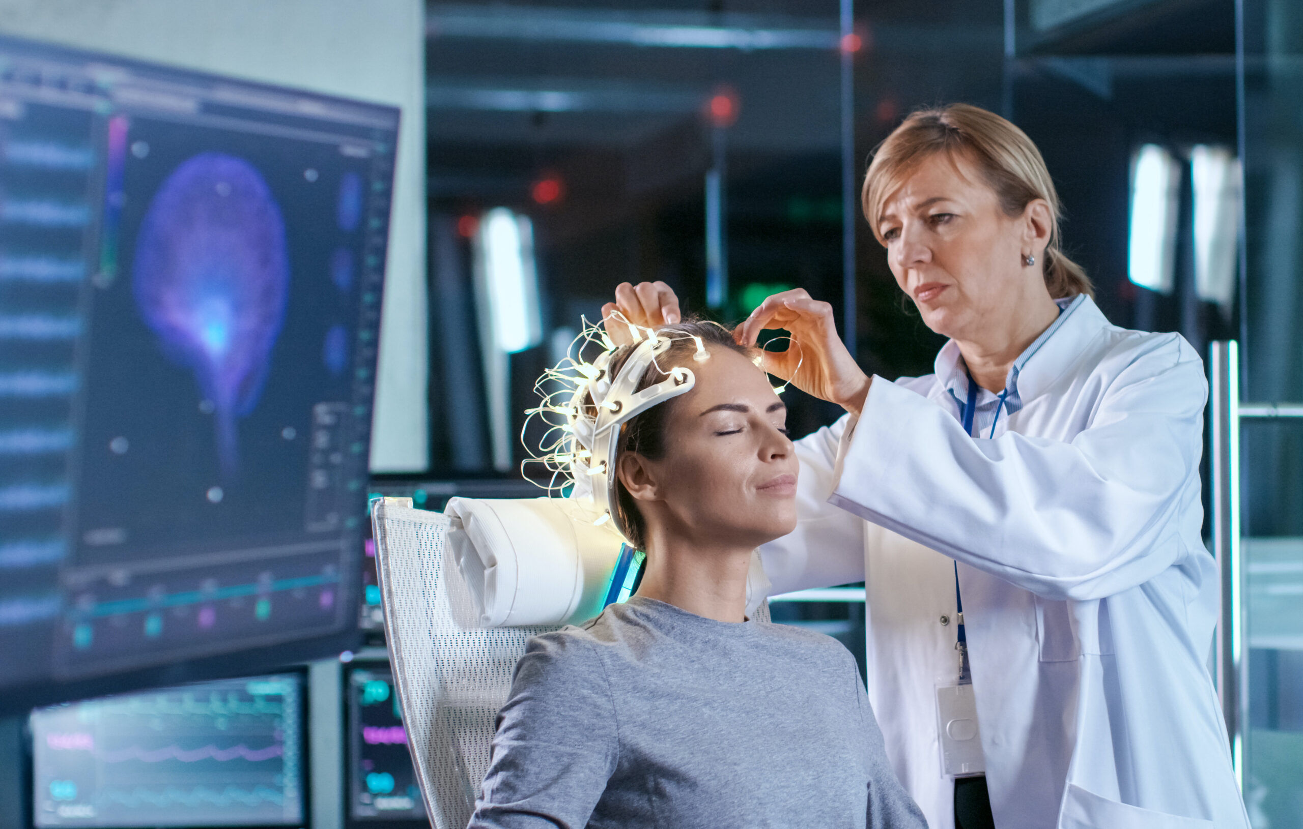 Woman Wearing Brainwave Scanning Headset Sits in a Chair while Scientist Adjusts the Device, Looks at Displays. In the Modern Brain Study Laboratory Monitors Show EEG Reading and Brain Model.