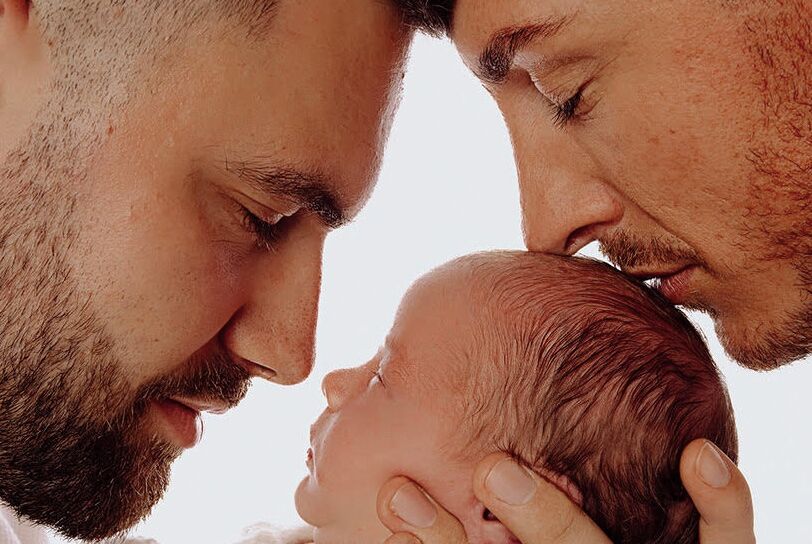 Two bearded men nuzzle their infant child in this promotional image