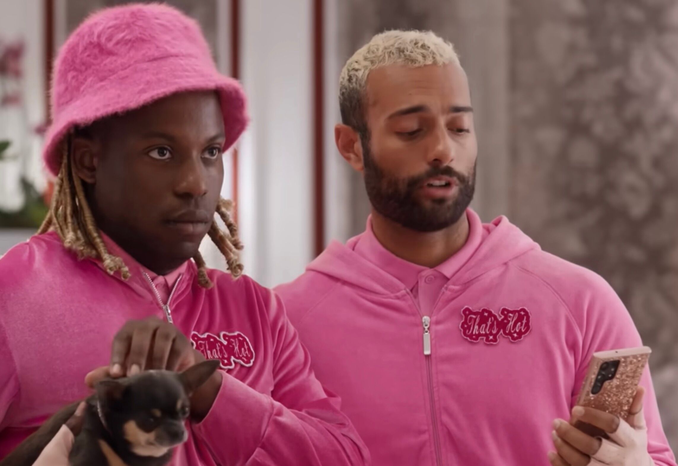 Two male presenting folks wearing pink, one holding a Chihuahua and the other texting