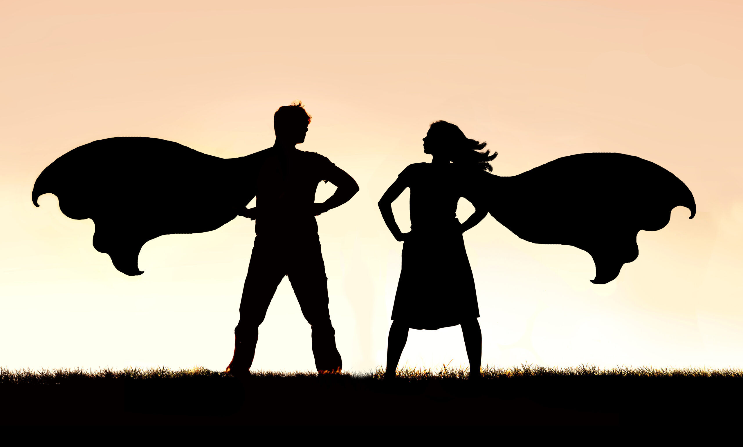 A silhouette of a superhero man and woman couple in capes standing strong and powerful against a sunset sky background.