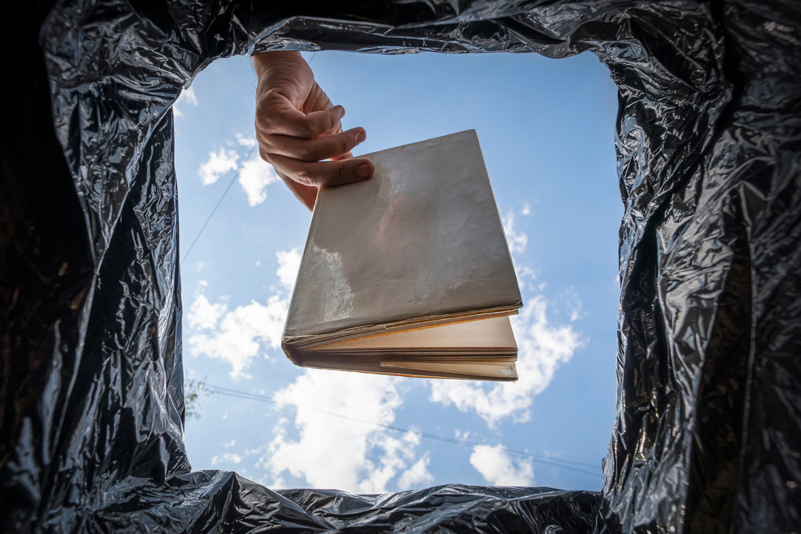 View from inside trash can as someone throws away a book against a sunny sky backdrop