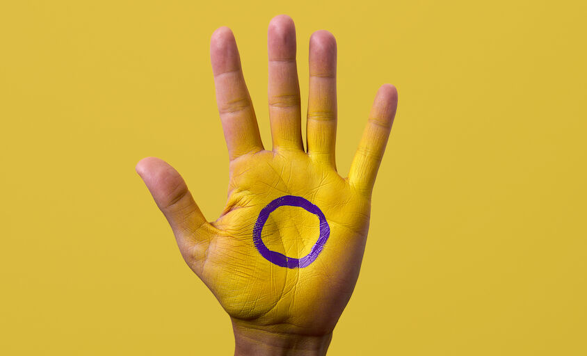 closeup of the intersex flag painted in the palm of the hand of a young person, against a yellow background with some blank space around it