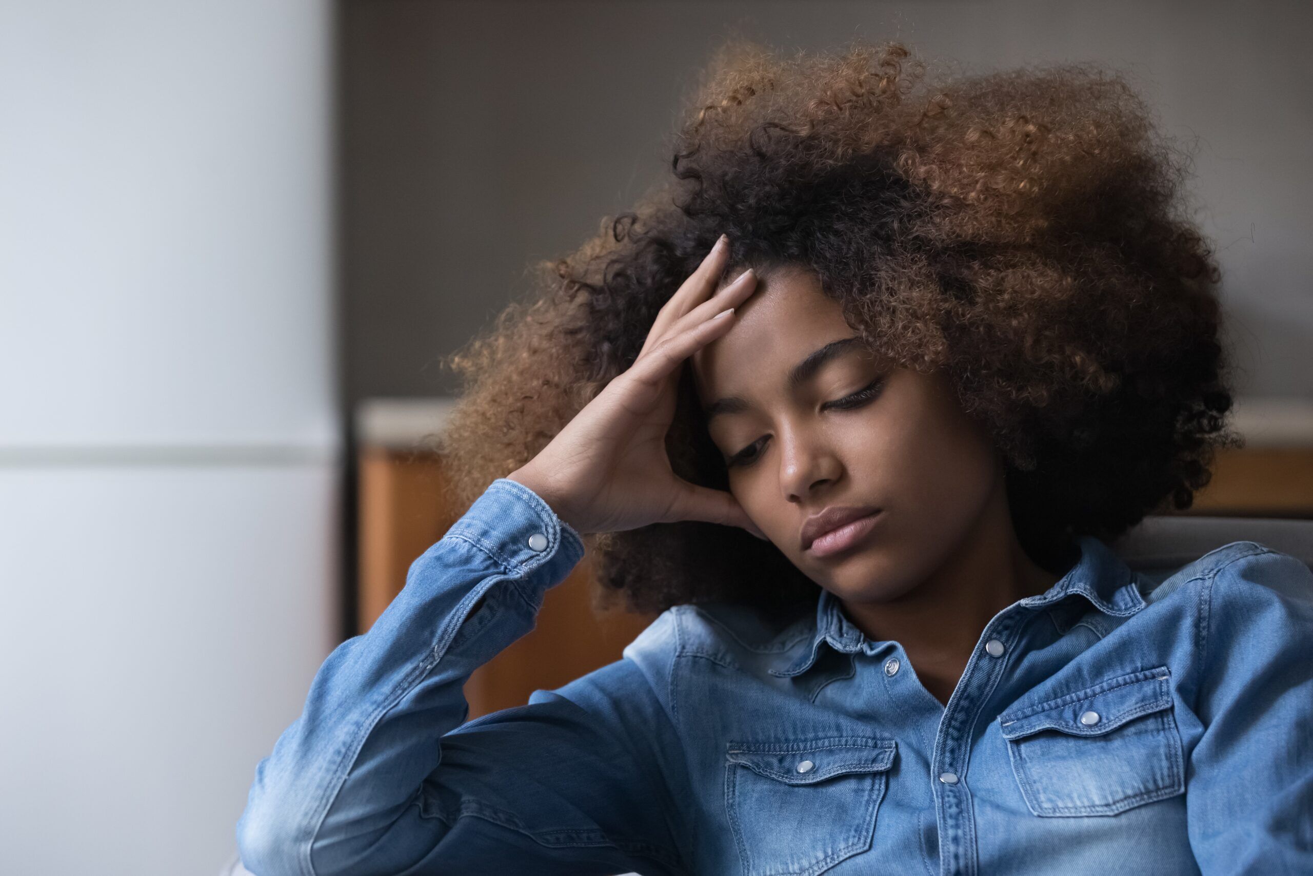 Frustrated unhappy African teen girl suffering from depression, melancholy at home, sitting on couch, touching head, feeling bad due to stress, hurt, despair, emotional problems, youth crisis