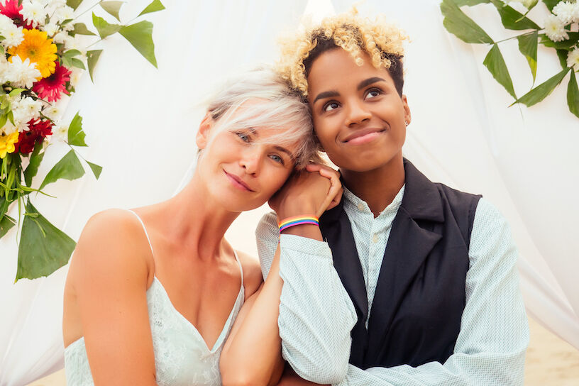 african american female groom in black suit and caucasian bride in dress in ceremony on tropical beach under wedding flover arch