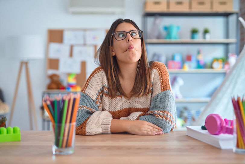 Young beautiful teacher woman wearing sweater and glasses sitting on desk at kindergarten making fish face with lips, crazy and comical gesture. Funny expression.