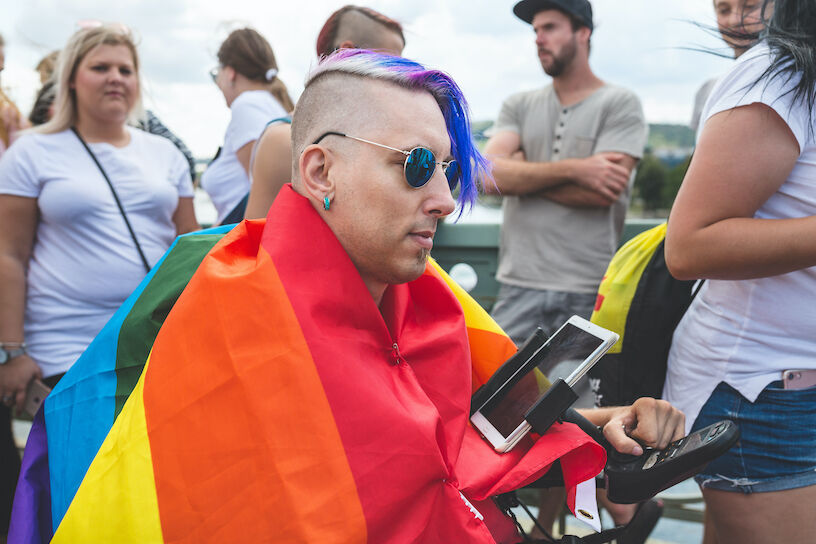 Prague, Czech Republic - August 11, 2018: People at the annual Prague Gay Pride parade. Hipster man in a rainbow flag in a wheelchair with a tablet in a crowd.