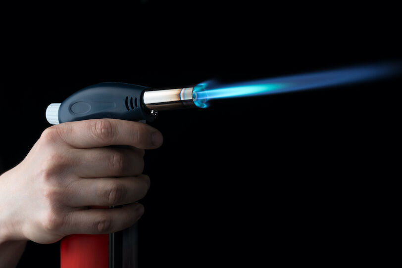 A white hand holds a blowtorch as a blue flame shoots from its metal nozzle.