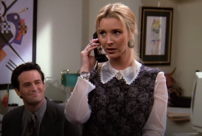 Chandler Bing, a white man in his late twenties, watches as his friend, Phoebe, a white woman with blonde hair, answers a phone as his personal assistant