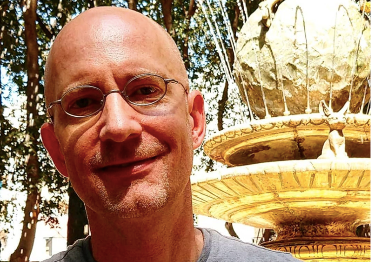 The author Michael Jensen is a bald middle-aged white man wearing glasses glasses and a white shirt standing in front of a yellow and green outdoor fountain in the sunlight