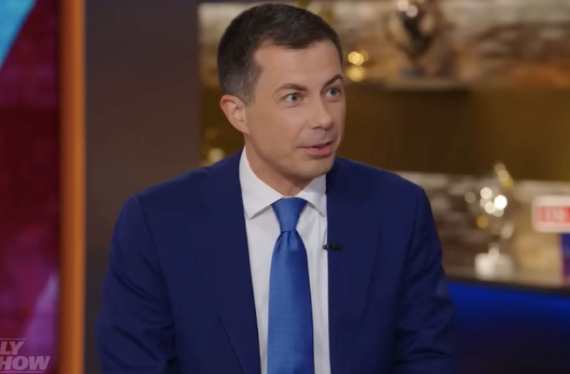 Pete Buttigieg, a white 42-year-old man, wears a blue blazer and tie while on the brown colored set of the TV news program.