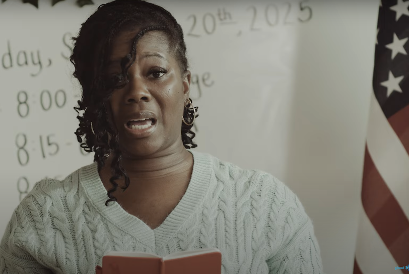 An image from GLAAD's ad on Project 2025 shows a Black teacher looking nervously as she recites an altered version of the Pledge of Allegiance in front of a whiteboard and a US flag