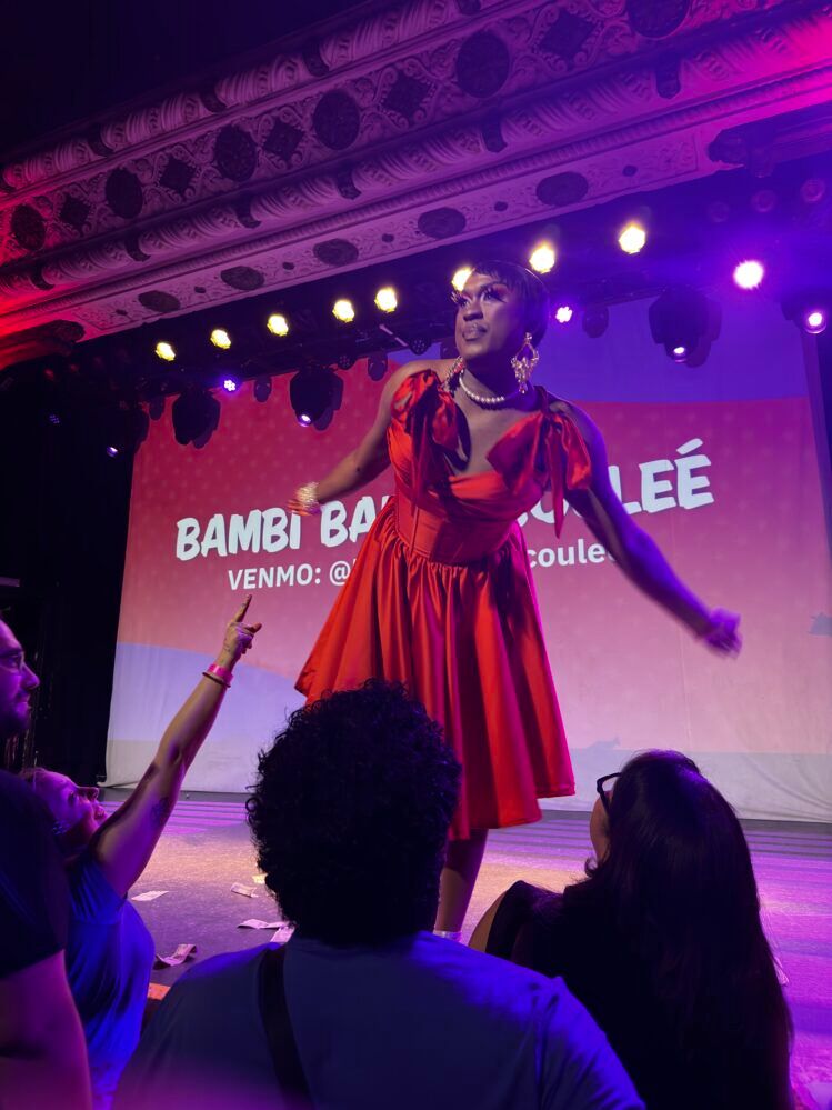 Bambi Banks-Coulee performing on stage at the Drag PAC event in Chicago. She is wearing a vibrant red dress with a plunging neckline, accessorized with pearl earrings and a pearl necklace.