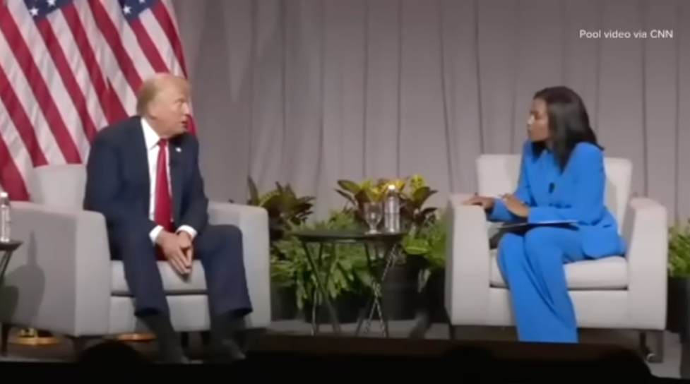 Trump wears a blue suit and his trademark red tie while sitting on a cushioned chair in front of an American flag onstage across from a black woman in a similar chair wearing a blue pant suit