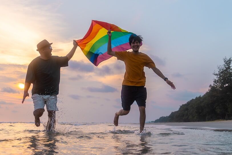 Happy Gay couple with lgbt flag running together on the beach in summer day. Happy guys demonstrate their rights. LGBTQI, Pride Event, LGBT Pride Month, Gay Pride Symbol.