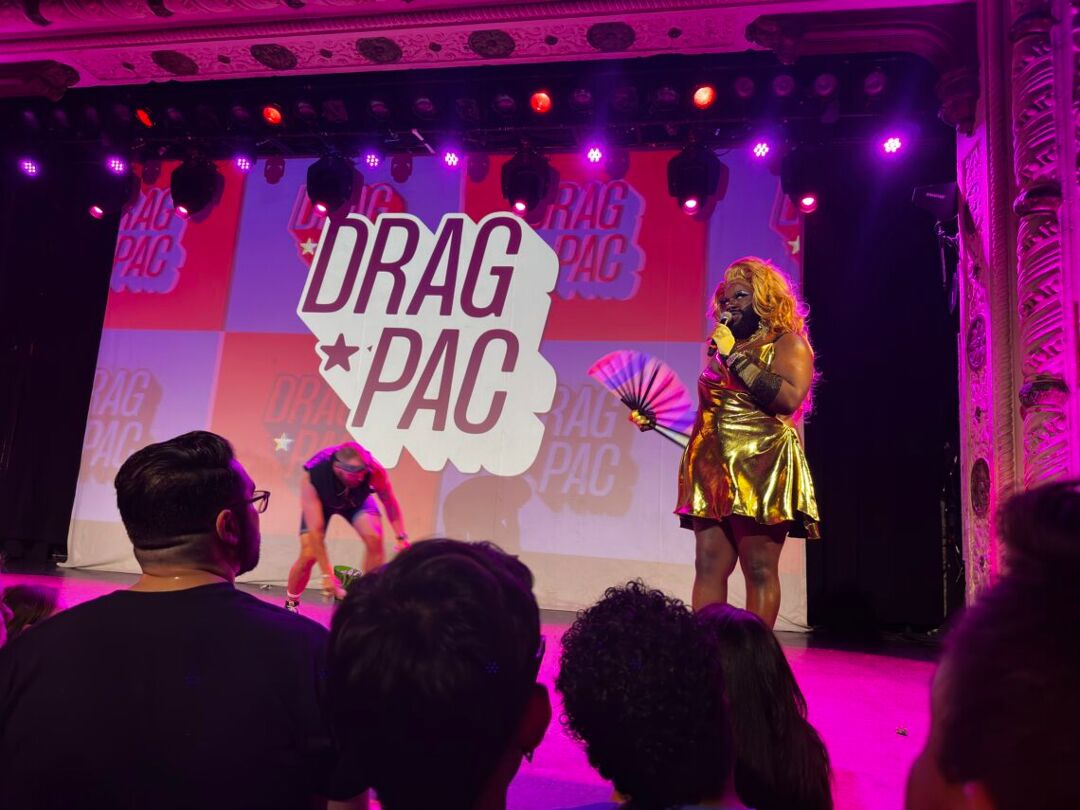 Host Lucy Stoole standing on stage in a gold dress with a rainbow clack fan in front of a Drag PAC logo at Drag Night Chicago