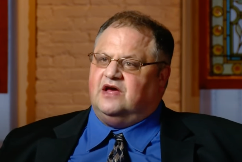 Steve Silberman is an older white man around age 60 something. In this photo, the white cleancut man wears a black blazer, blue collared shirt, and black tie while sitting in a wood-paneled room for an interview.