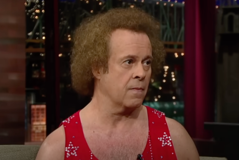 Richard Simmons is a white man with curly reddish brown hair wearing a red tank top shirt with silver stars on it.