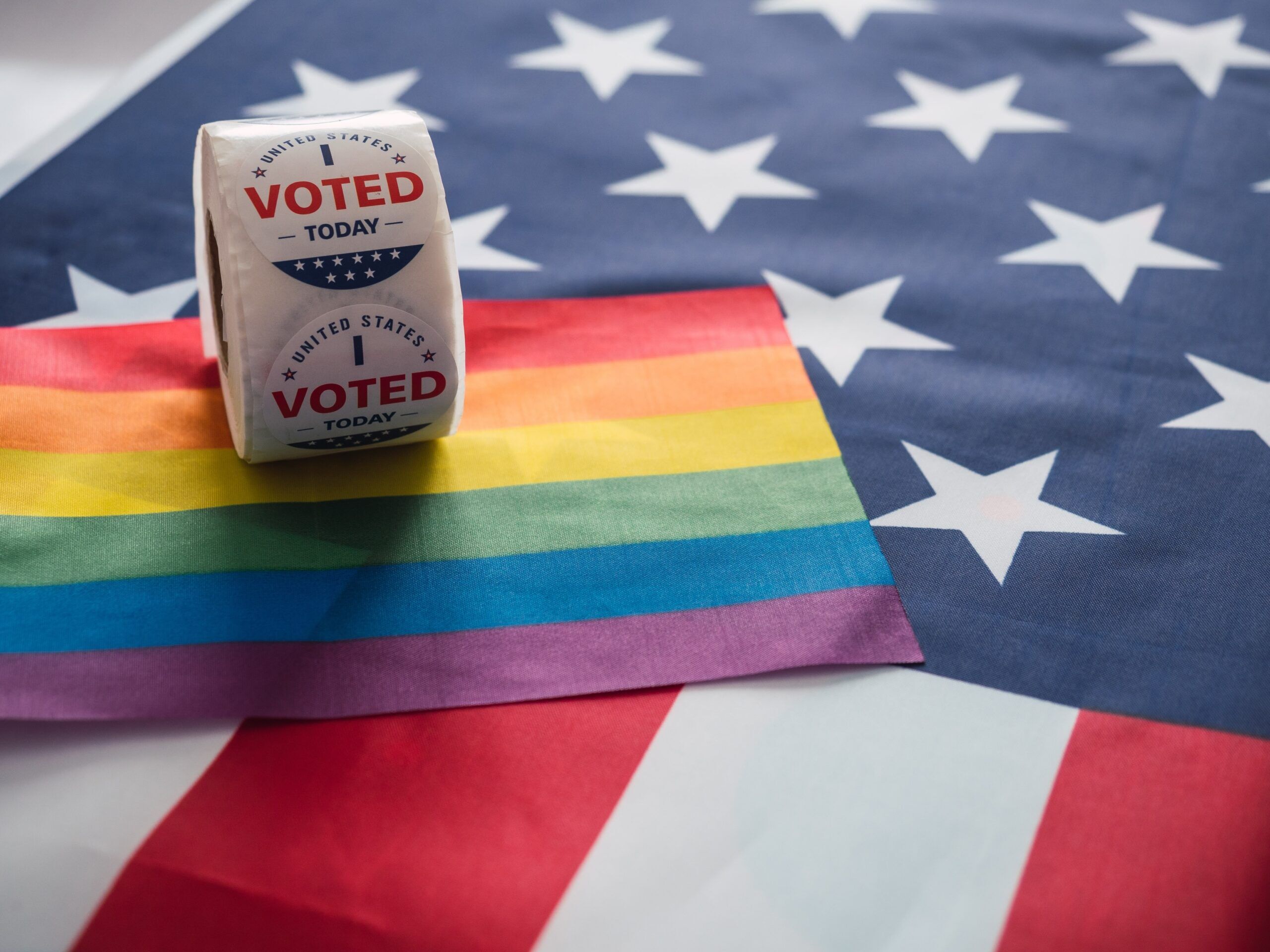 A roll of “I Voted Today” stickers is placed on a rainbow pride flag, which is laid over an American flag. The image symbolizes the intersection of LGBTQ+ pride and civic duty in the United States, with the vibrant colors of the rainbow flag contrasting against the stars and stripes of the American flag.