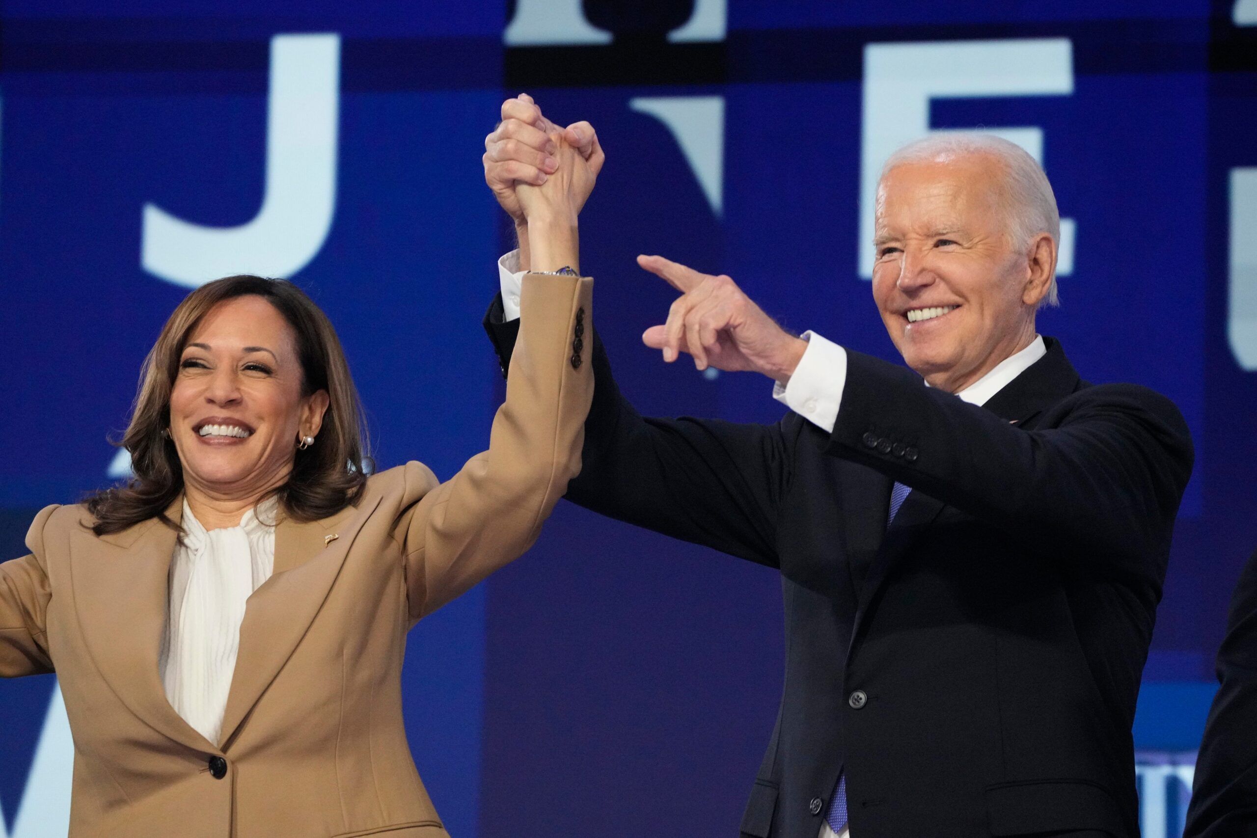 Vice President Kamala Harris and President Joe Biden stand on stage, holding hands in a celebratory gesture. Both are smiling brightly; Kamala Harris is wearing a beige suit, while Joe Biden is dressed in a dark suit with a white shirt. The background features a blue and white design.