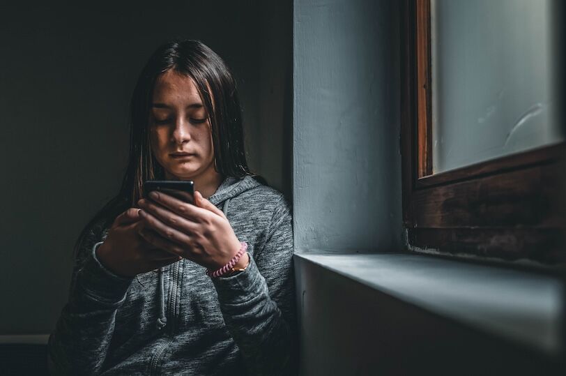 Teenage girl using smart phone in the dark. Sad teenager with mobile phone, scared of threatening, mobile abuse. Front view of a sad teen checking phone