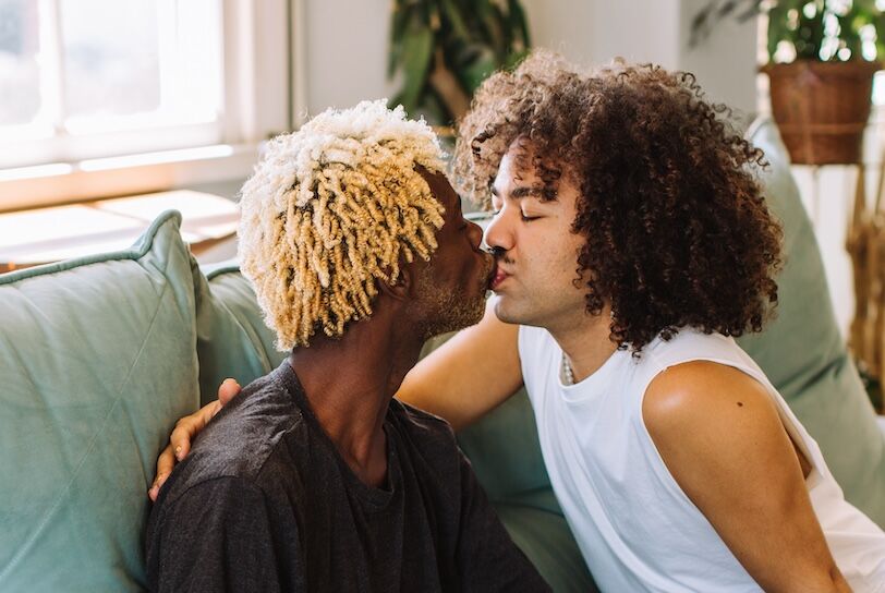 Passionate young gay couple being romantic indoors. Two affectionate male lovers kissing each other while sitting together in their living room at home. Young gay couple in love.