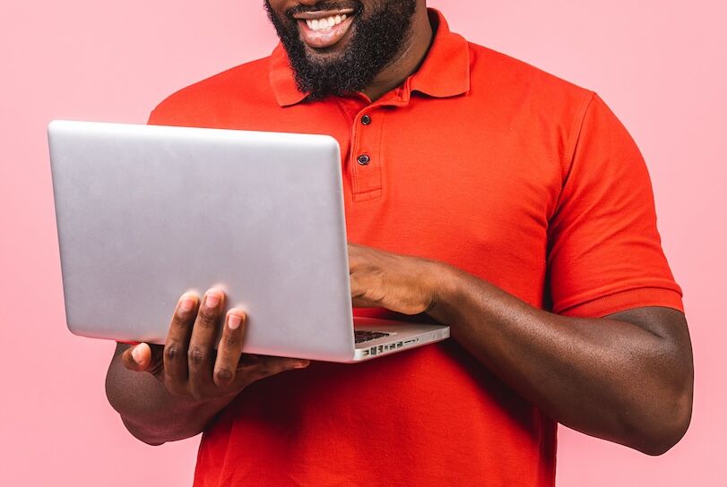 Young smiling african american man standing and using laptop computer isolated over pink background.