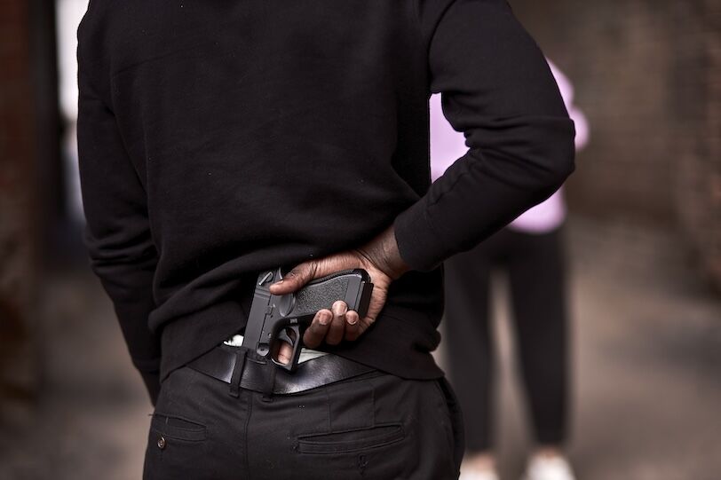 Robbery or criminal man in black hoodie is holding gun standing behind the woman, going to commit a crime. view from back on male in black outfit, close-up hands. criminal activity concept