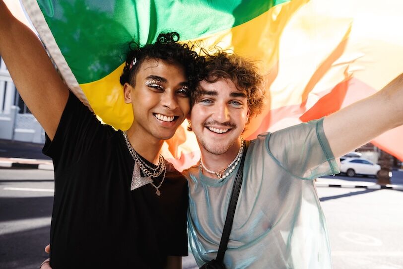 Two young queer men celebrating gay pride. Two young gay men smiling cheerfully while raising a rainbow flag at a gay pride parade. Two members of the LGBTQ+ community standing together outdoors.