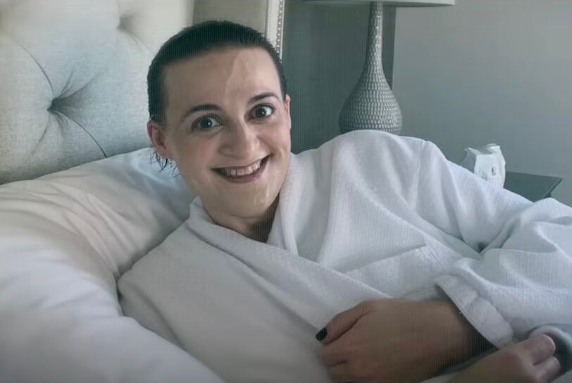 Elisabeth Finch lays in a white bed smiling while clutching a robe around her.