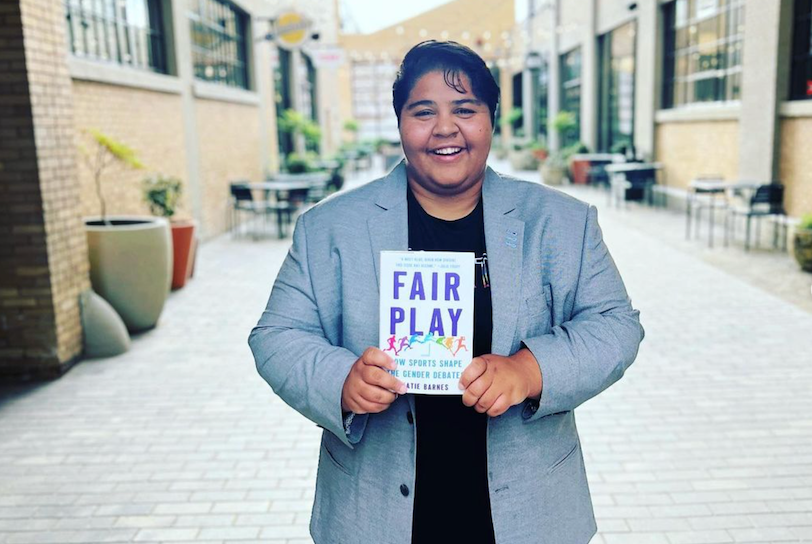 Katie Barnes is a nonbinary journalist with tanned skin and brunette hair. They stand ourdoors, in a corridor of white brick buildings and hold a copy of their book, entitled "Fair Play: How Sportrs Shape the Gender Debate"