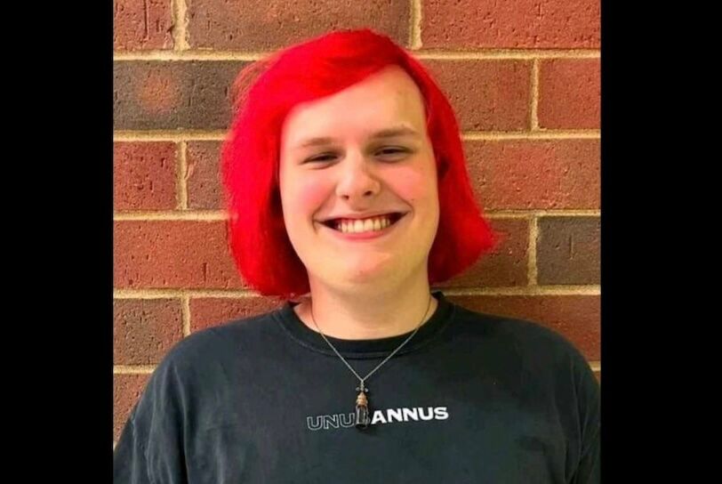 Serenity Birdsong is a white transgender young person with red hair smiling against a brick wall.