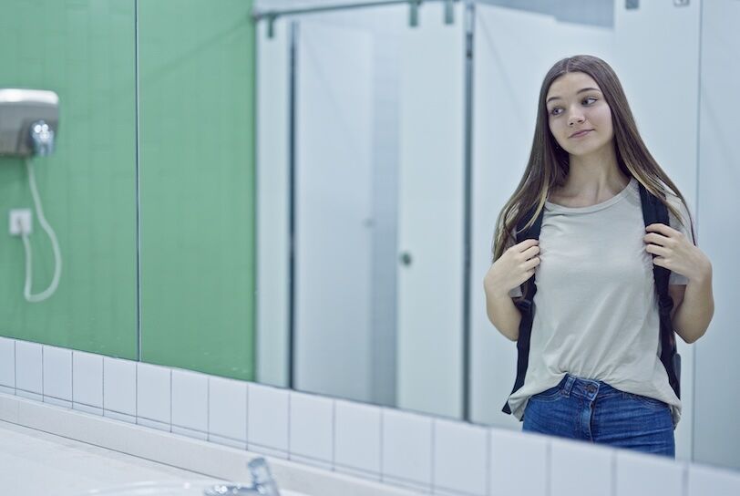 Young beautiful girl student wearing backpack looking on mirror at bathroom