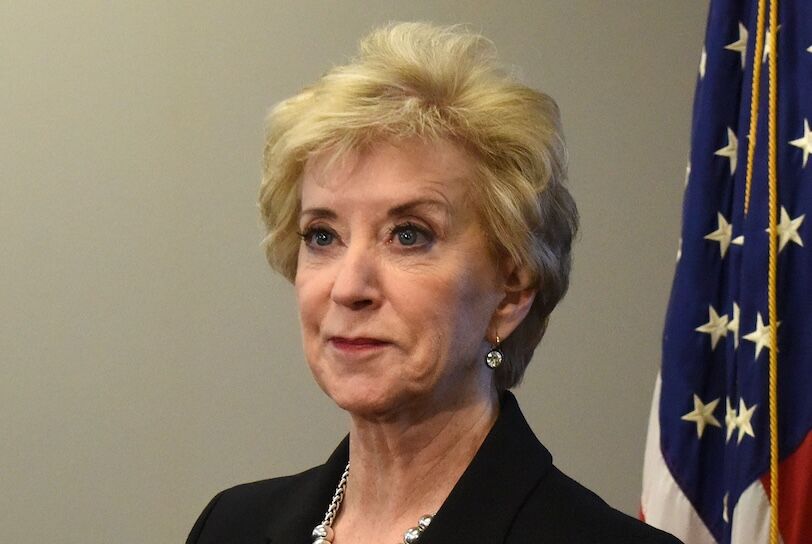 Linda McMahon is a blonde old white woman wearing a necklace and black blazer in front of a U.S. flag.