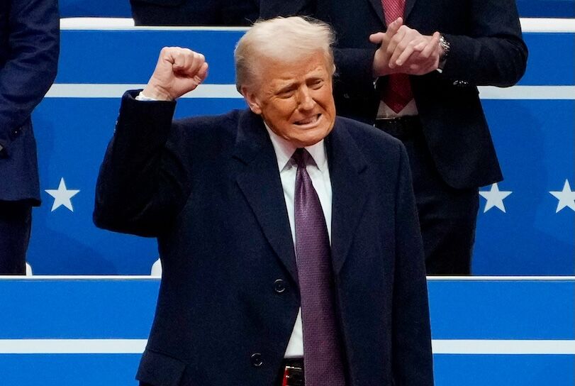 Donald Trump, a 78 year old white man with a pale yellow combover, raises his fist while wearing a black overcoat and tie. He stands on a dais with a blue background and white stars.