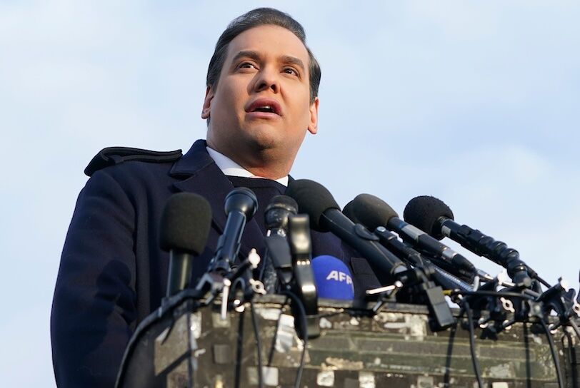George Santos is a white brunette guy speaking outdoors at a lectern filled with microphones.